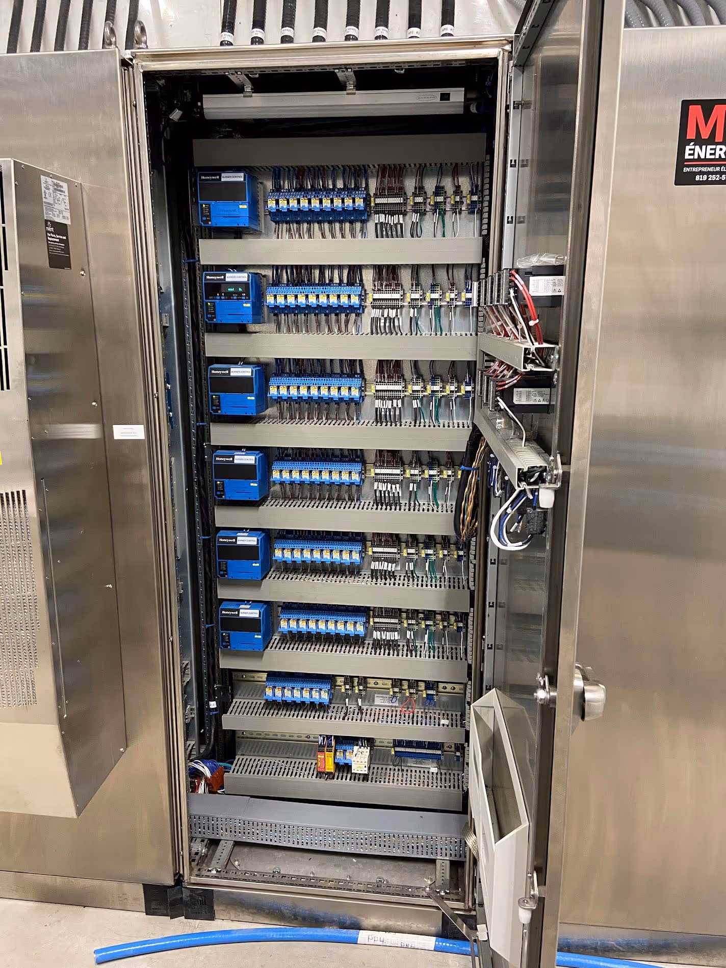 Armoire électrique industrielle ouverte montrant des rangées ordonnées de composants tels que des disjoncteurs, relais, connecteurs et câblage organisé à l'intérieur d'un boîtier en métal inoxydable.