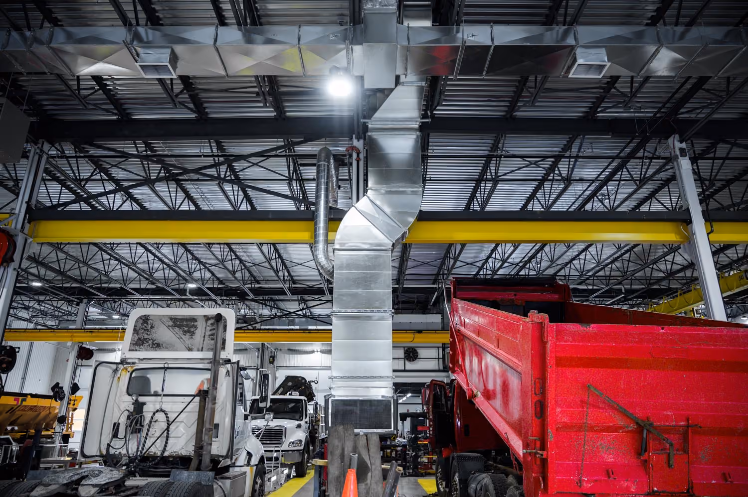 Vue intérieure d'un atelier mécanique avec plusieurs camions en réparation, dont un camion rouge à benne basculante à droite et un camion blanc à gauche, sous une grande structure de plafond en métal avec des conduits d'aération et des poutres jaunes.