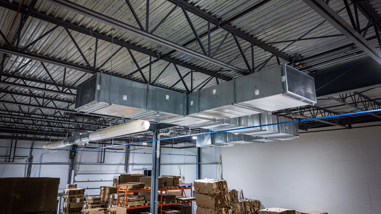 Vue intérieure d'un entrepôt avec des conduits métalliques suspendus au plafond en acier, et des palettes empilées contenant des cartons et matériaux divers.