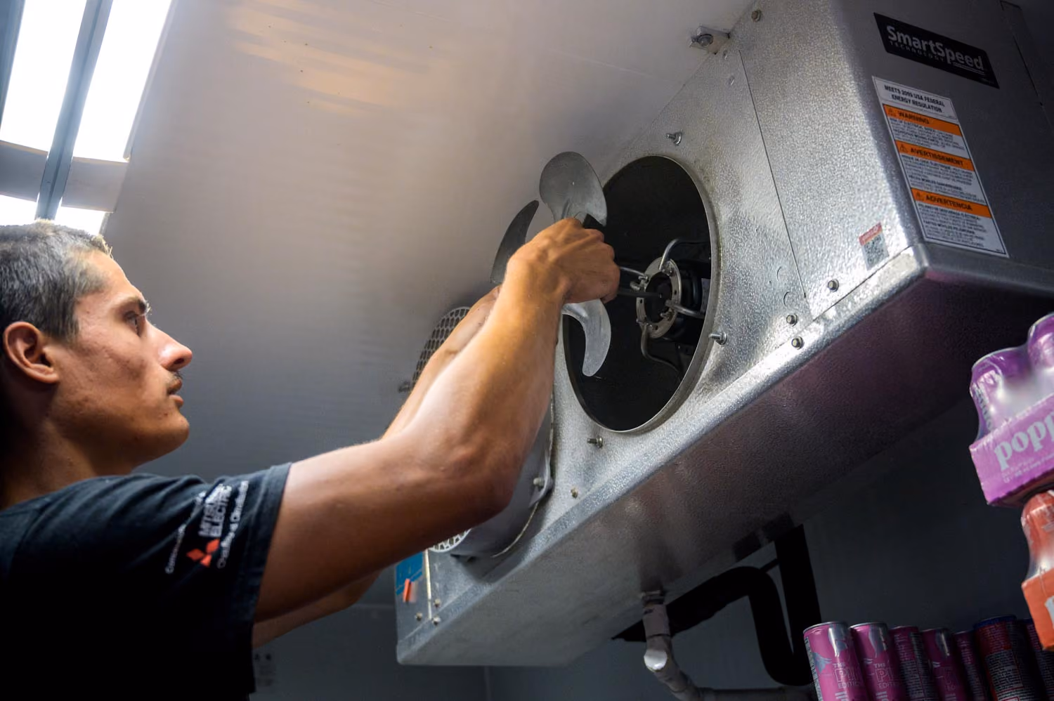 Un homme réparant un ventilateur industriel mural, tenant une pale en métal devant l'ouverture du ventilateur dans une pièce avec faible éclairage et canettes empilées à proximité.