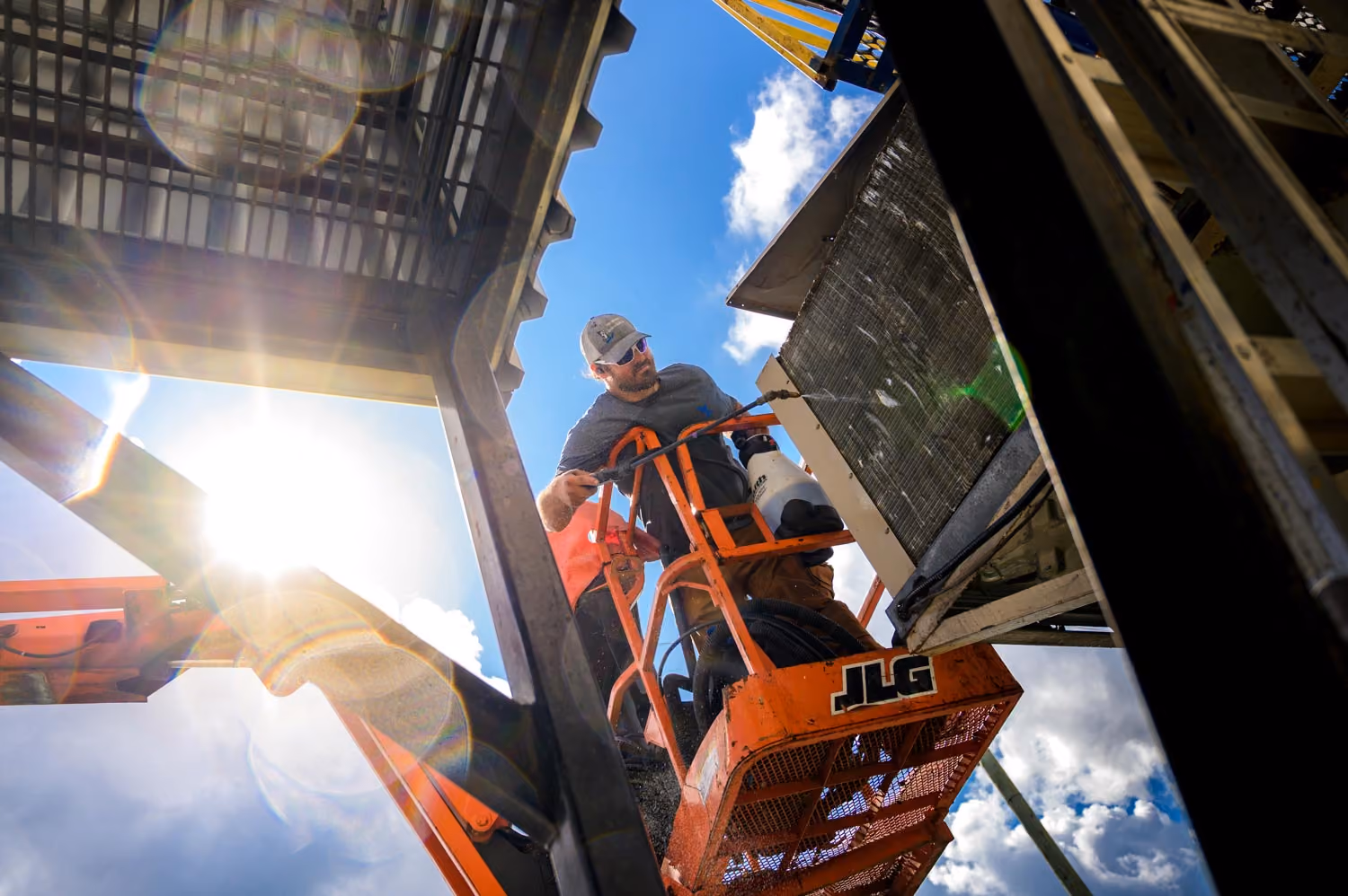 Un ouvrier vêtu d'une casquette et de lunettes de soleil travaille sur une plateforme élévatrice orange JLG sous un ciel bleu avec quelques nuages, manipulant un équipement industriel.