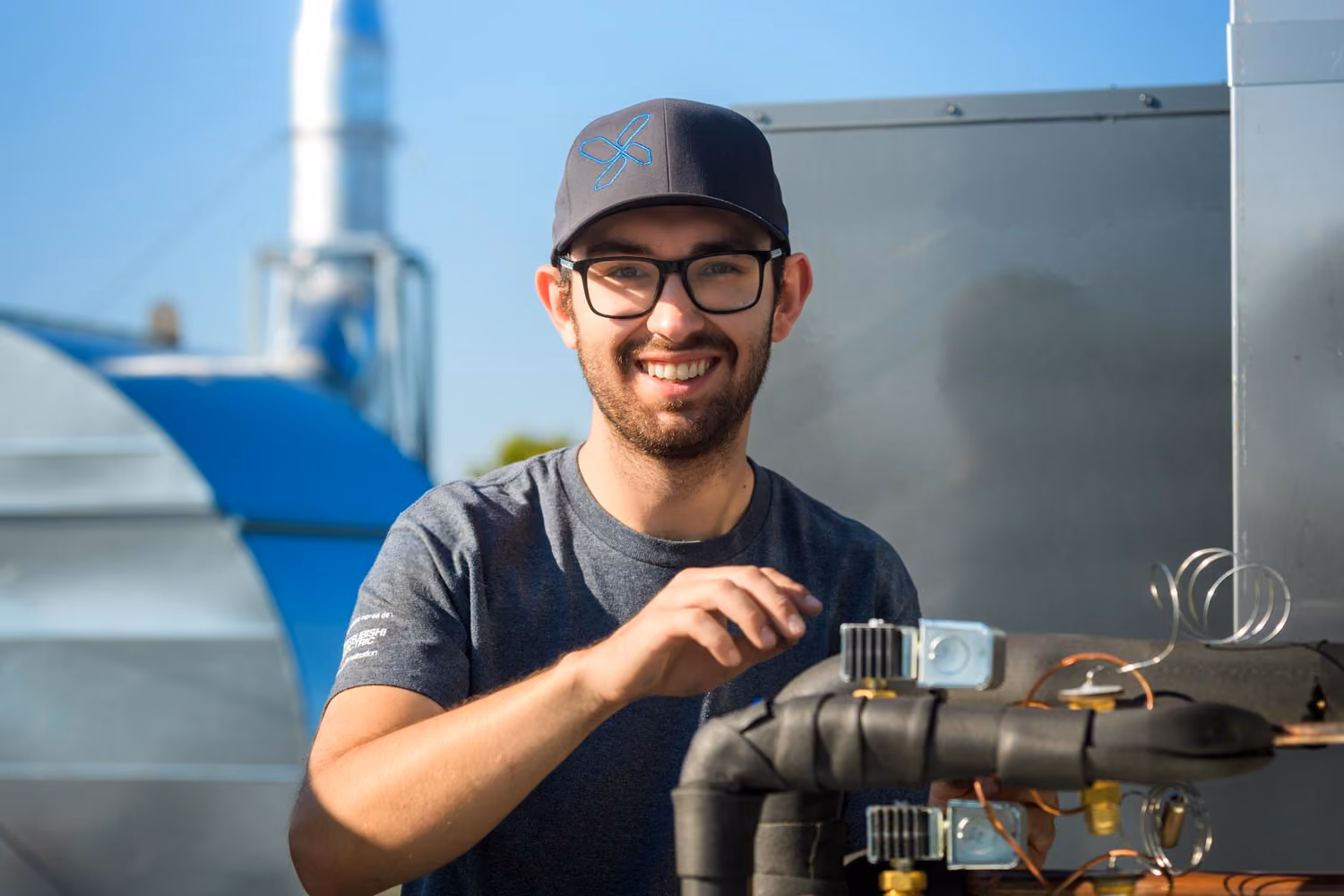 Un homme souriant portant une casquette noire et des lunettes, vêtu d'un t-shirt gris, travaille sur un équipement technique isolé à l'extérieur par une journée ensoleillée.