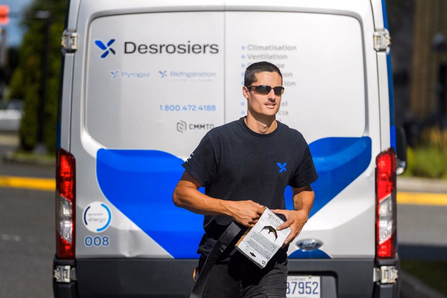 Un homme en t-shirt noir portant des lunettes de soleil tient un dispositif et se tient devant une fourgonnette blanche et bleue portant le logo et le nom « Desrosiers ».