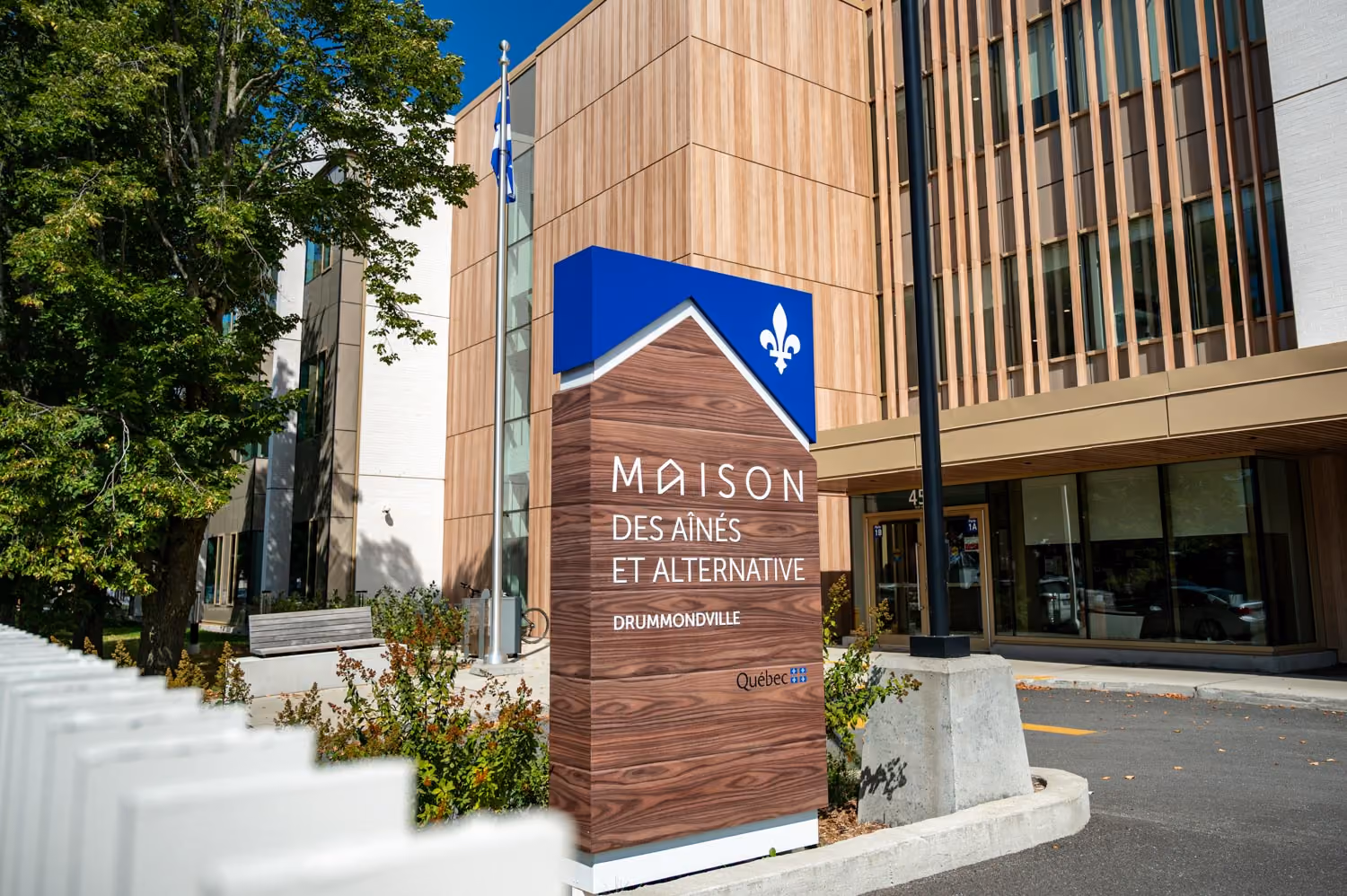 Panneau de la Maison des Aînés et Alternative à Drummondville, Québec, à l'entrée d'un bâtiment avec des murs en bois clair et une fenêtre vitrée, entouré d'arbres et d'une clôture blanche.