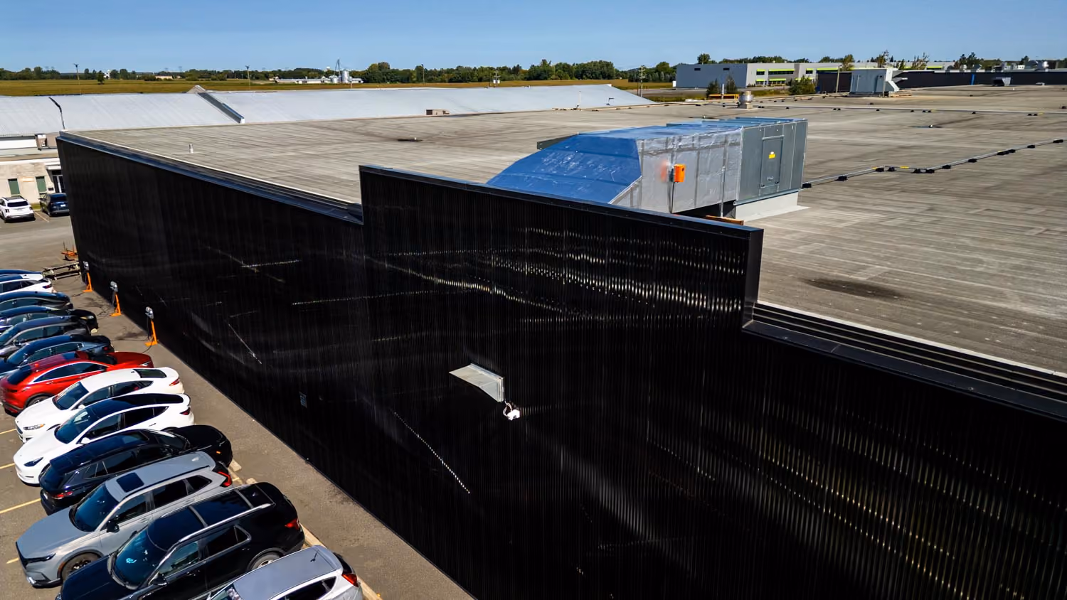 Vue aérienne d'un bâtiment industriel avec un mur extérieur noir et un toit plat, et une rangée de voitures stationnées à côté.