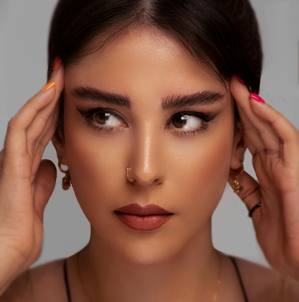 Close-up of a woman with bold eyebrows, winged eyeliner, and red lips touching her temples.