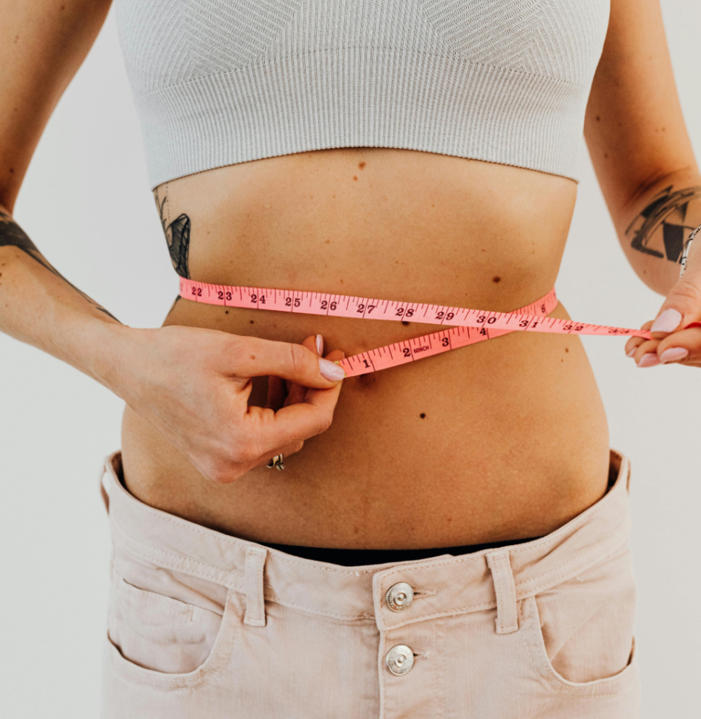Person measuring waist with pink tape measure while wearing a grey crop top and light pants.