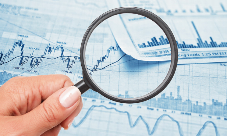 A hand holds a magnifying glass over a pile of paper with graphs and lines.