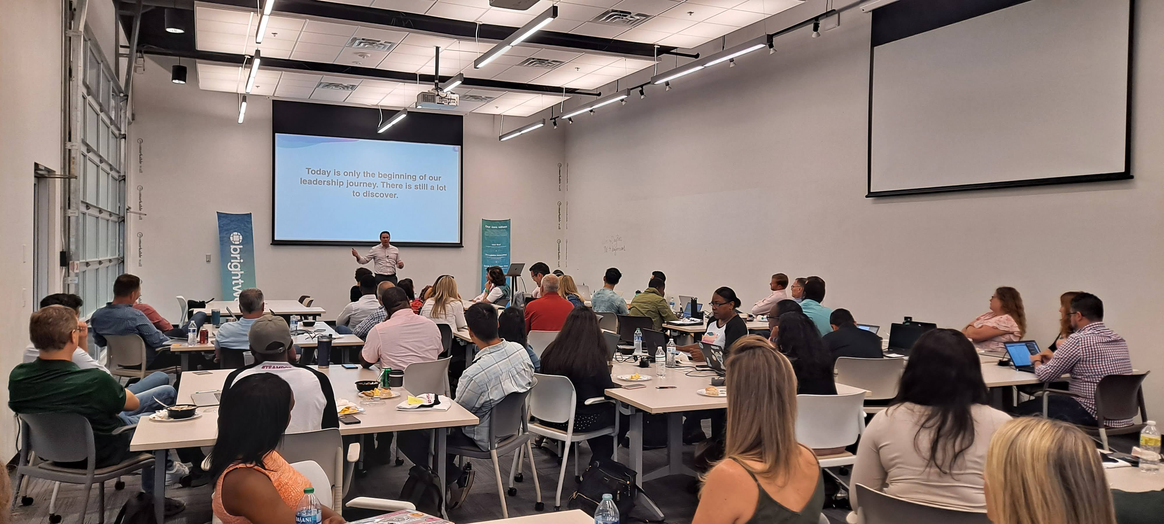 Groups of Brightwell employees sit pod-style at tables in a conference room while CEO Larry Hipp presents a PowerPoint at the front of the room. The fintech company's logo is displayed on a banner to the left of the room.