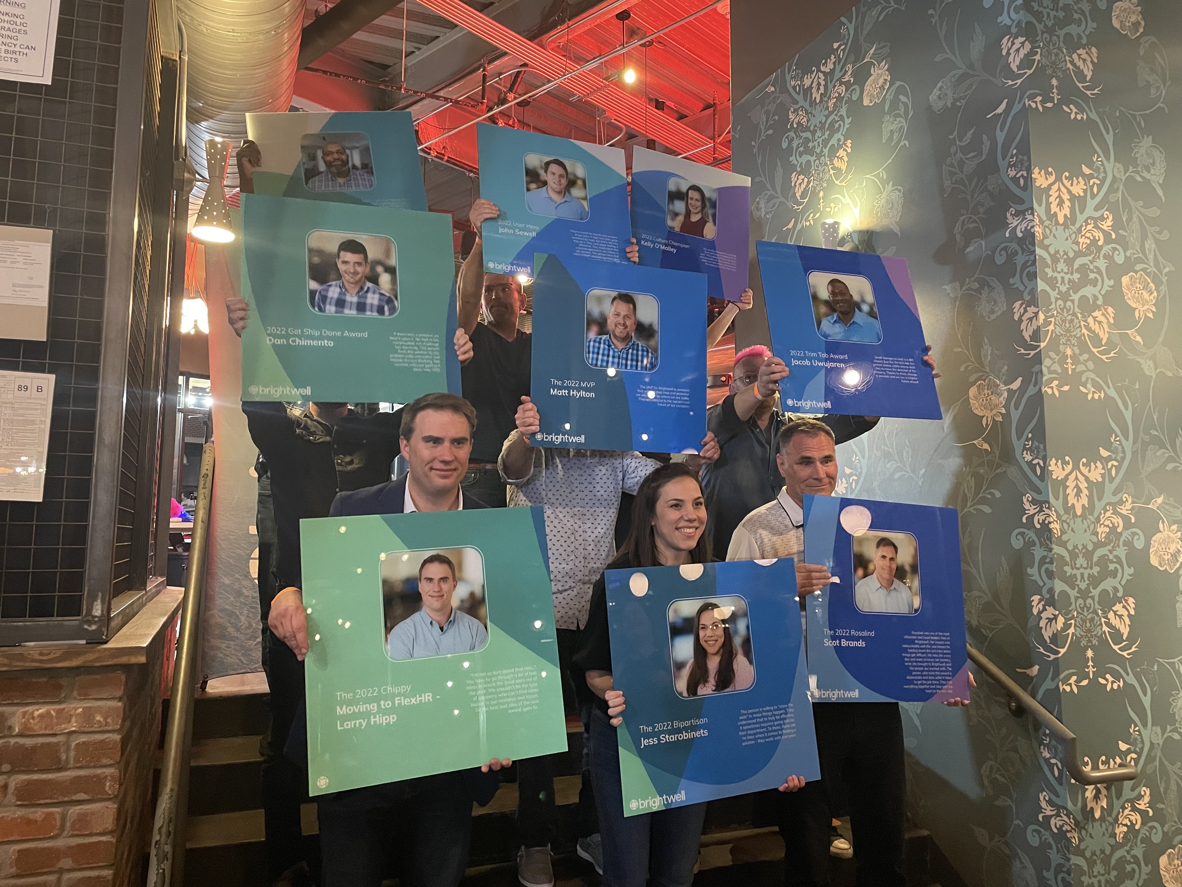 9 Brightwell employee award winners hold up their award posters which feature their name, award title, and description of the award category on a blue, green, teal, and purple branded background.