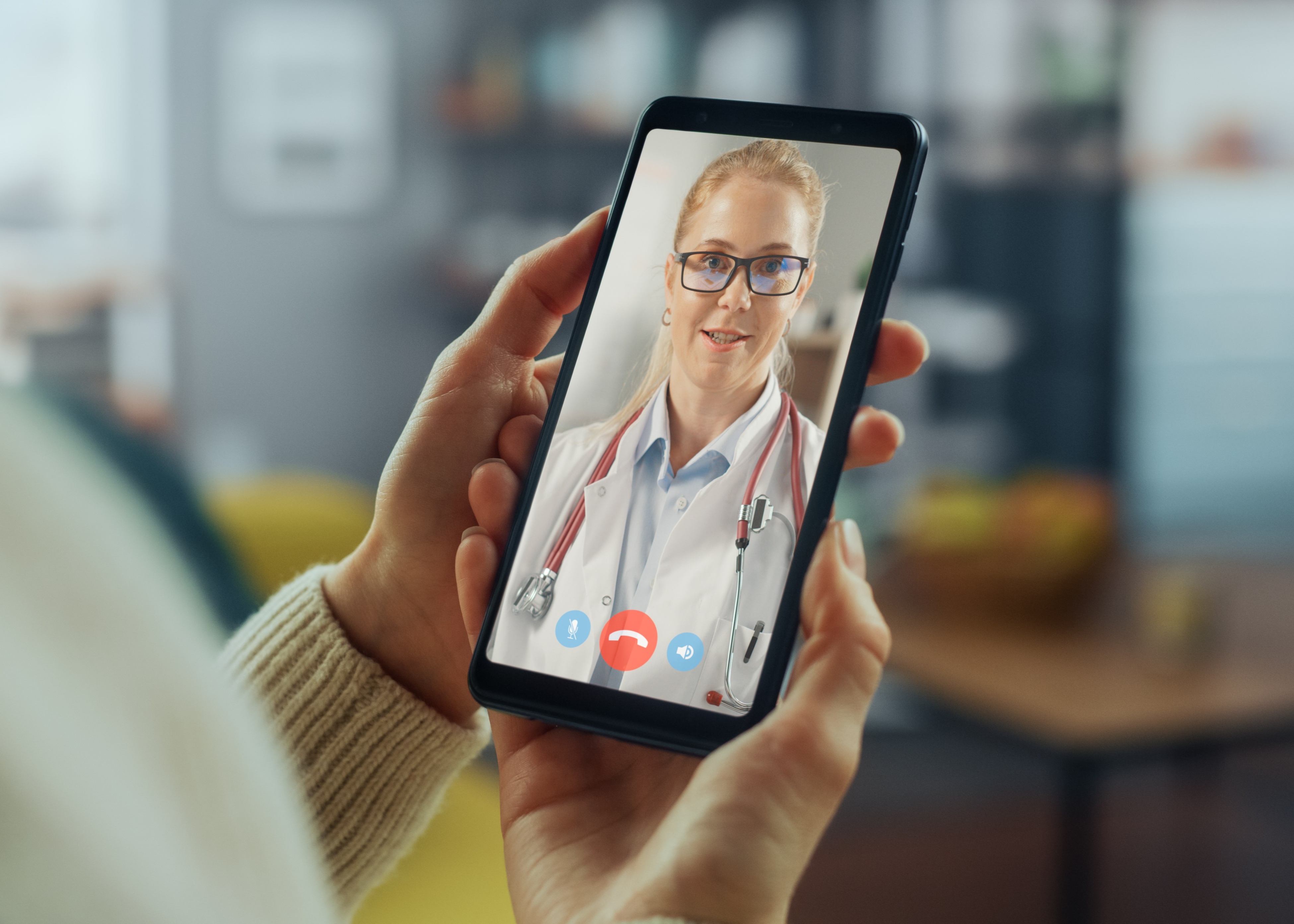 Person holding a smartphone showing a female doctor with glasses and a stethoscope during a video call.