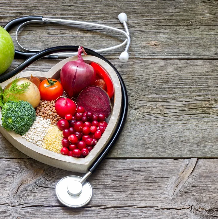 Heart-shaped wooden bowl filled with assorted healthy foods including onions, tomatoes, broccoli, grains, and berries, with a stethoscope resting on a wooden surface.