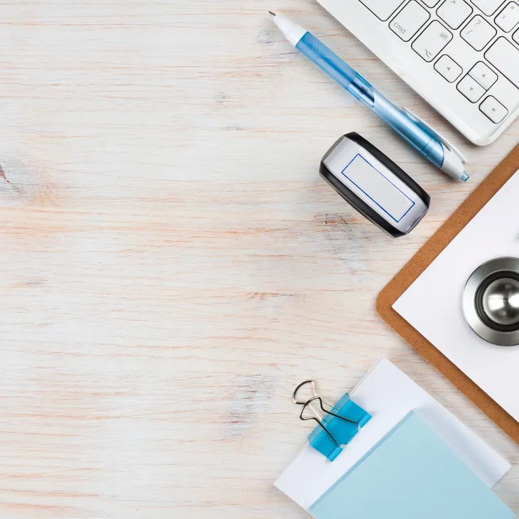 Workspace with a blue pen, stamp, keyboard corner, clipboard with stainless steel stethoscope, and blue binder clip on white wooden surface.