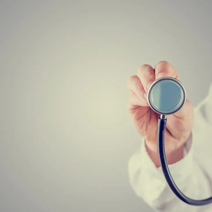 Hand holding a stethoscope chest piece towards the camera against a plain background.