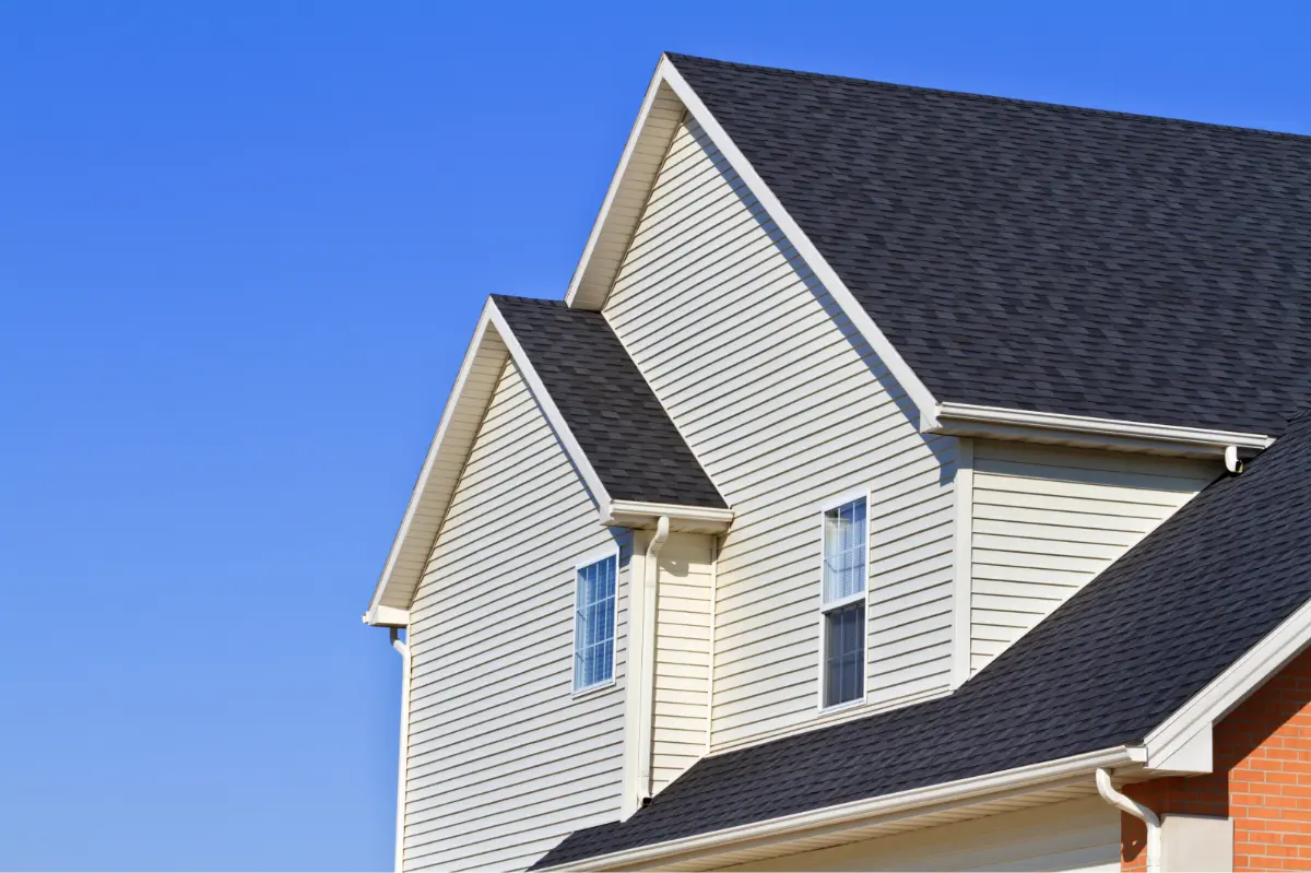 Upper portion of a beige siding house with black shingled gable roofs under a clear blue sky.
