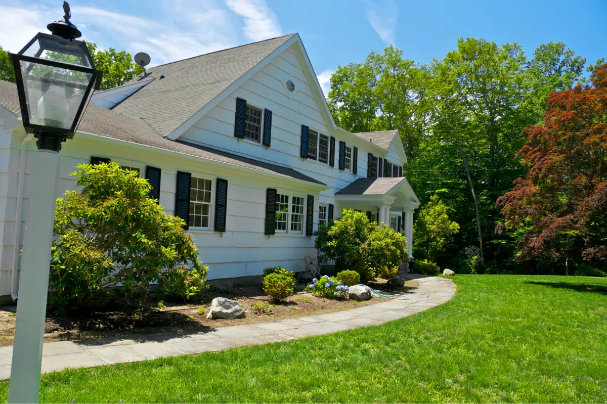 White two-story house with black shutters, surrounded by green bushes and a lawn under a blue sky.