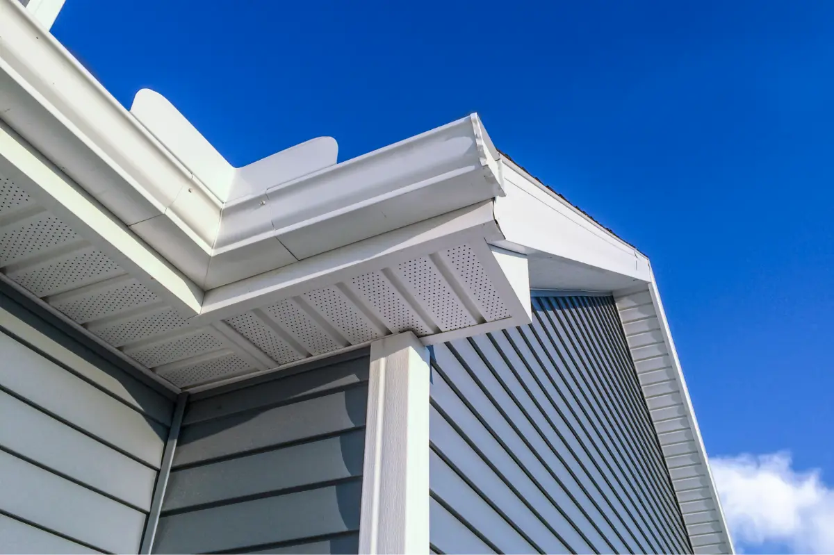 Close-up of a house corner showing white gutters, soffits, and gray siding under a clear blue sky.
