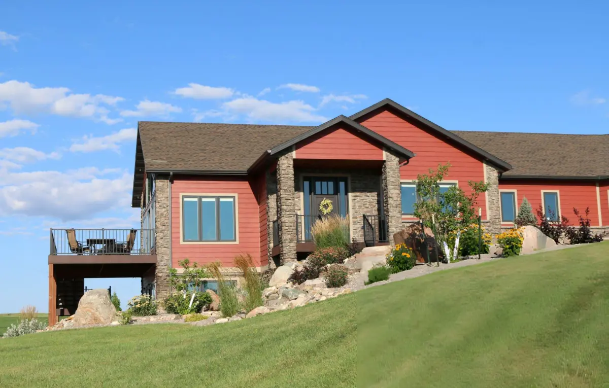 Red and stone house with a sloped roof, outdoor deck with chairs, and a green lawn under a blue sky.