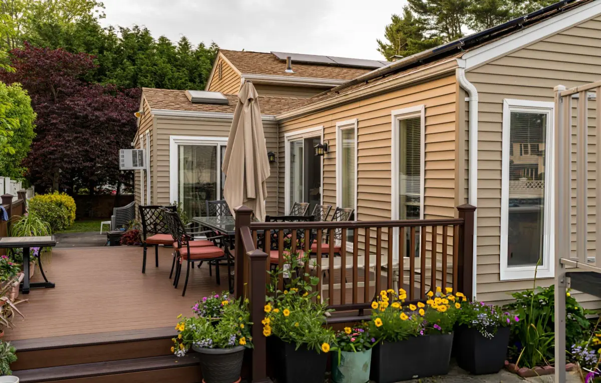 Backyard patio with brown wooden deck, outdoor dining table with chairs, beige closed umbrella, and flower pots with yellow and purple flowers.