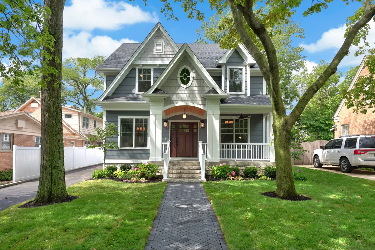 Two-story gray house with white trim, a central wooden front door, a stone pathway, green lawn, and large trees.