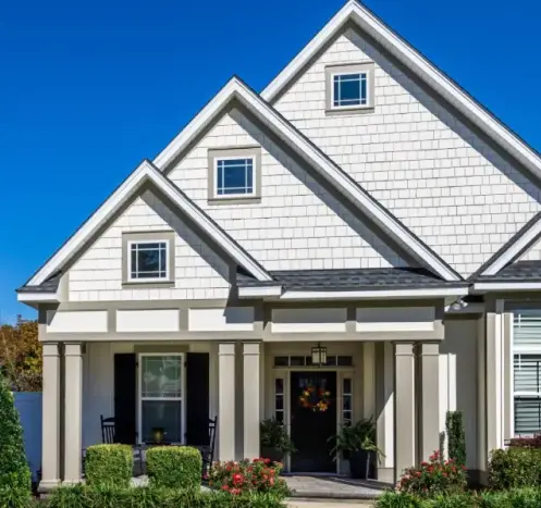 White house with three front gables, black door with fall wreath, and landscaped garden under a clear blue sky.