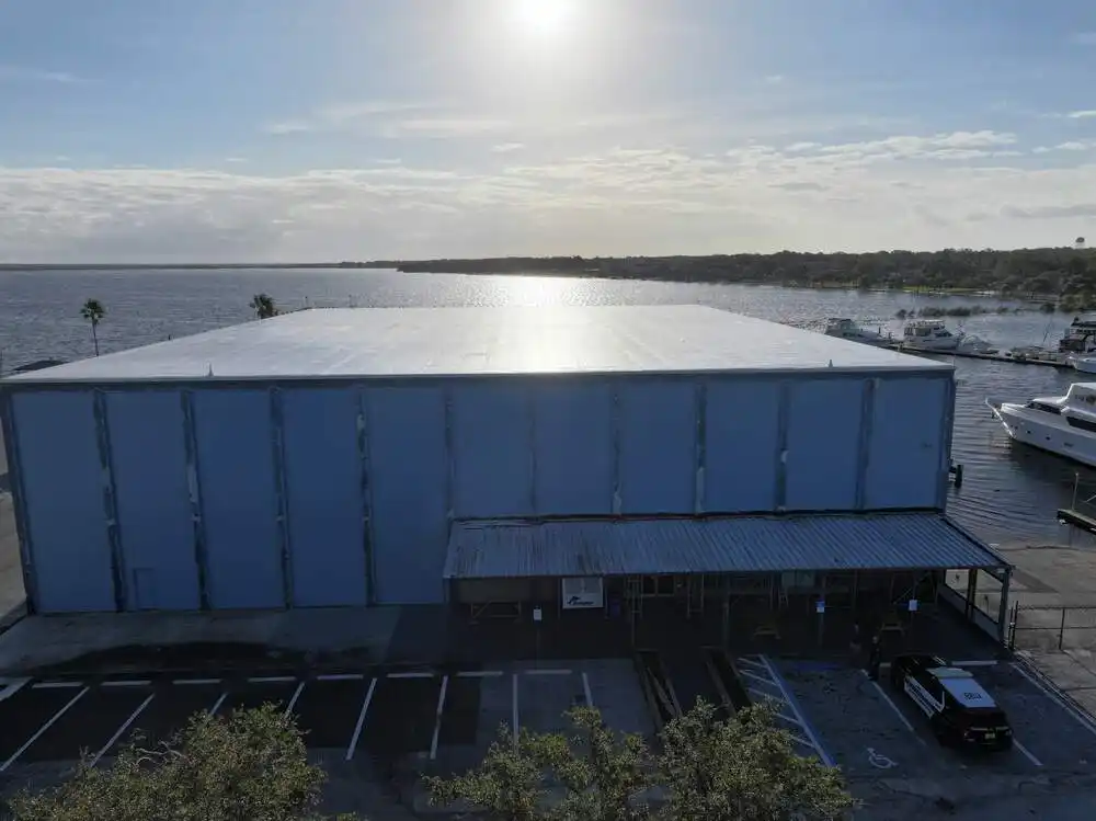 Large industrial building by water with boats docked and parking area in foreground under a bright sky.