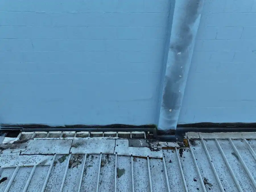 Weathered roof edge with damaged flashing and rust near a blue wall and drainpipe.