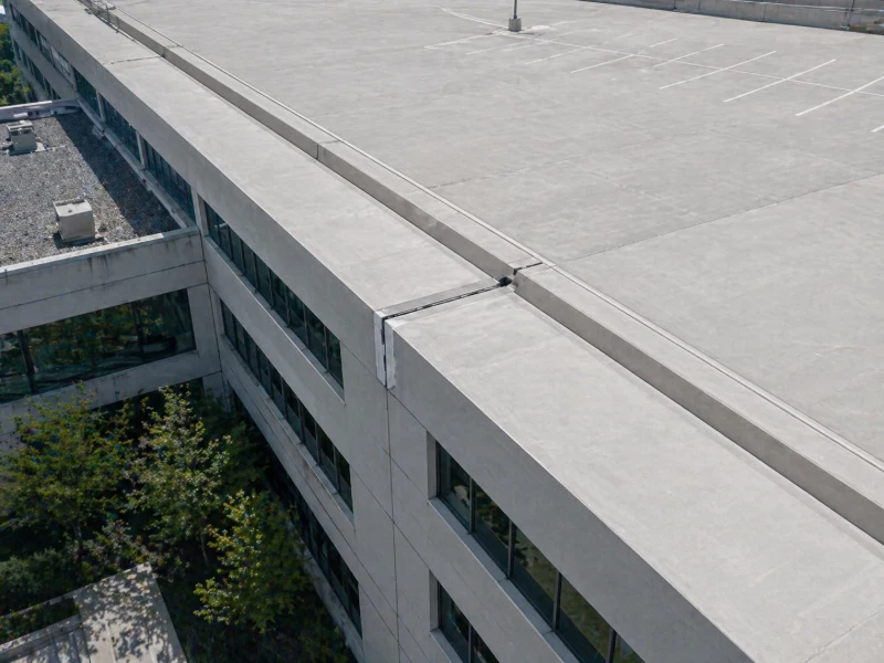 Concrete rooftop and parking lot of a multi-story office building with windows and some trees below.
