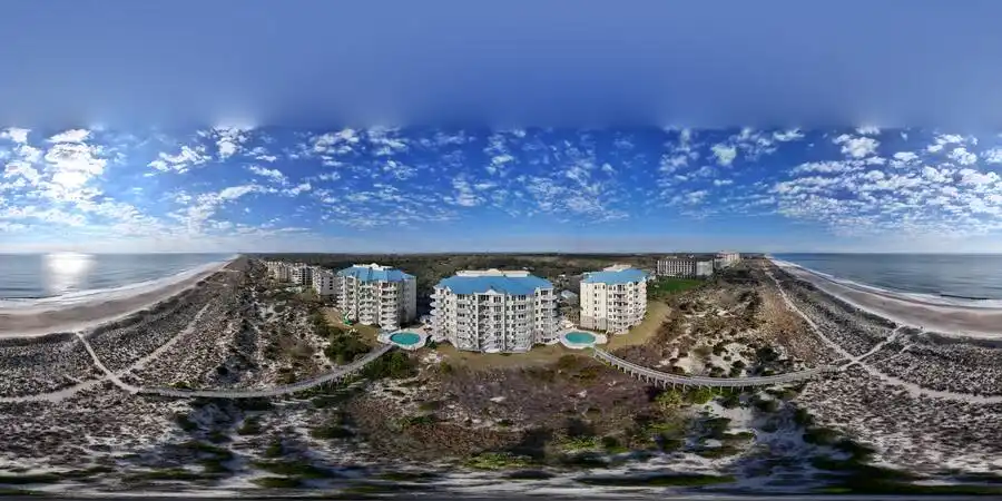Beachfront condos with blue roofs and pools surrounded by sand dunes under a blue sky with scattered clouds.