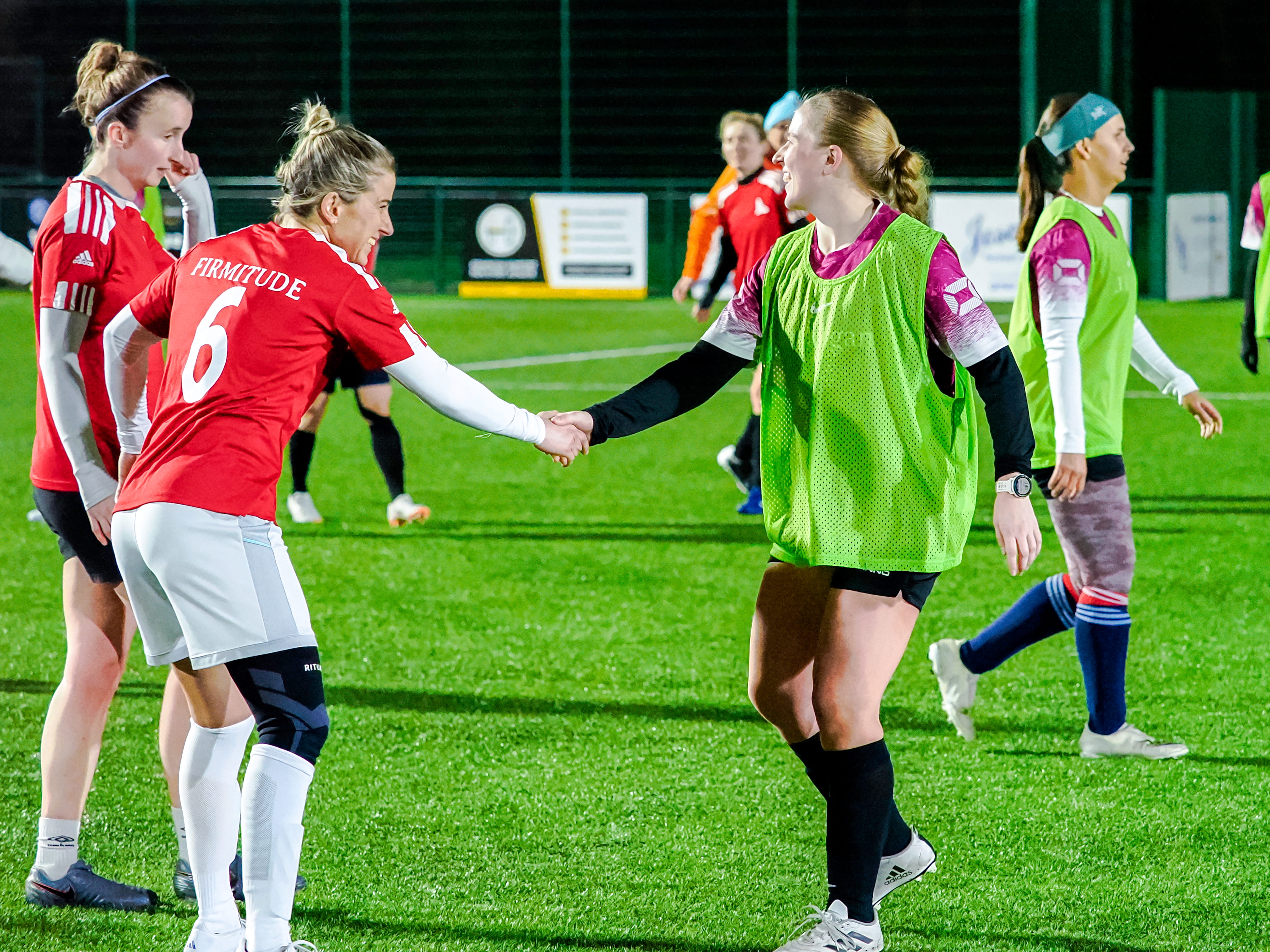 Two female football players shaking hands on a green field at night, one in a red jersey and the other in a green training vest.