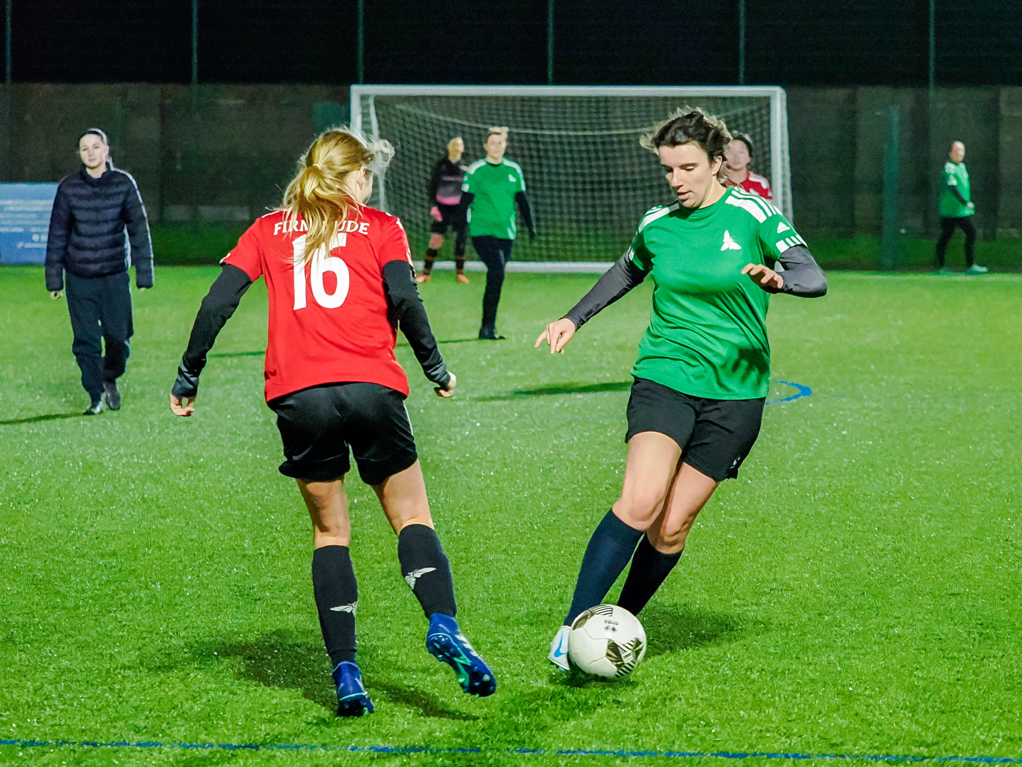 Two female football players competing for the ball on a brightly lit field at night, with a goal and other players visible in the background.