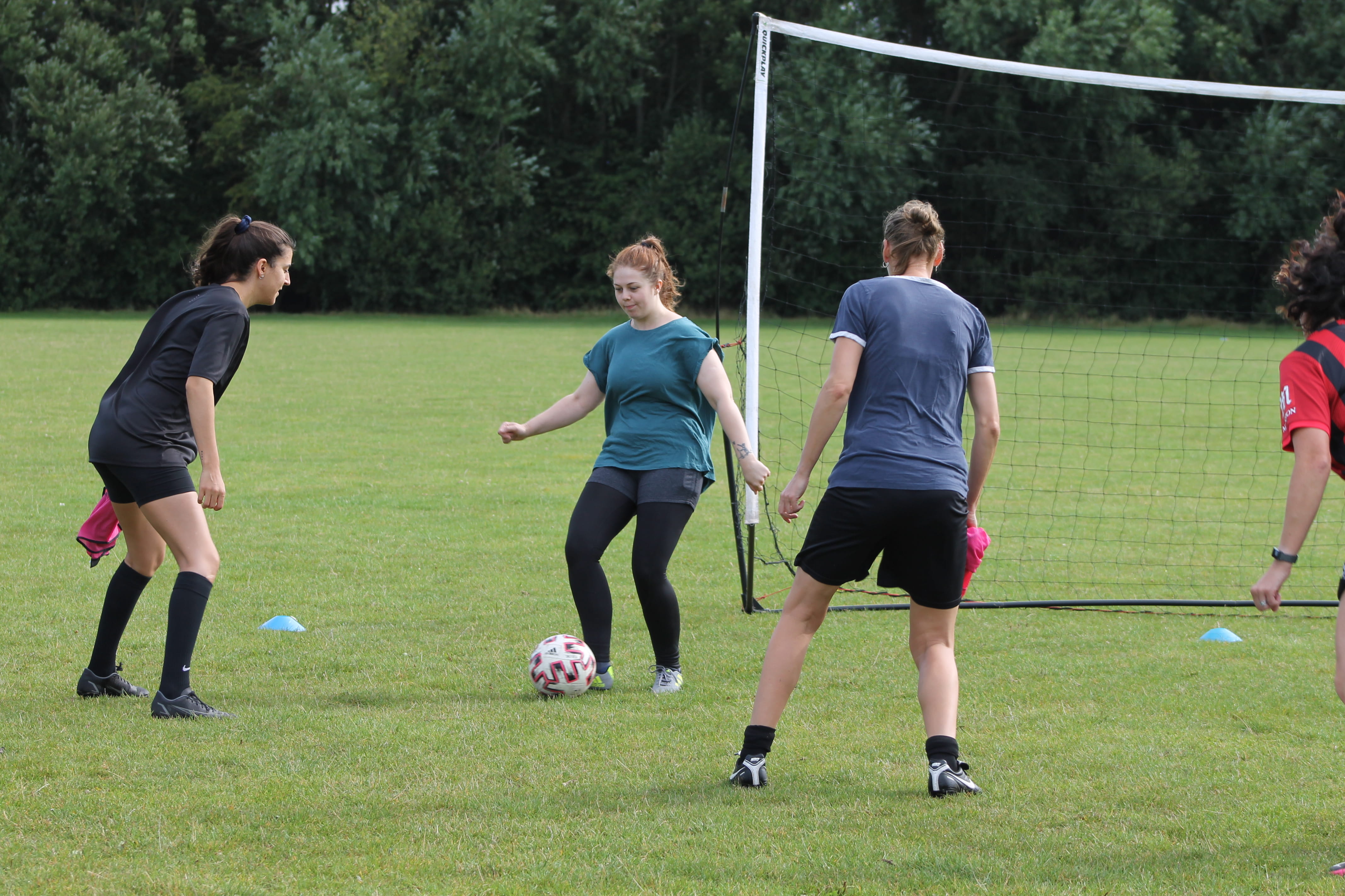 Four women playing football on a field near a goal net, one controlling the ball.