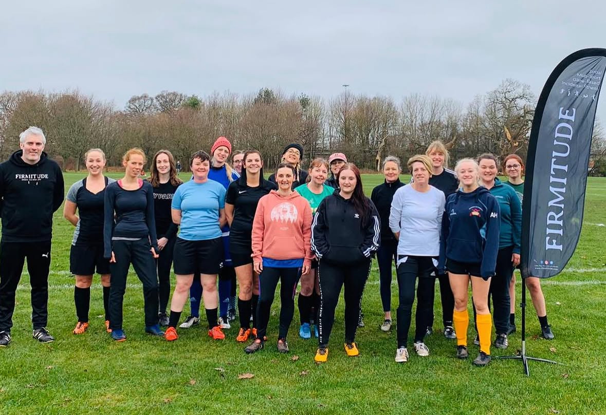 Group of women and one man standing on grass field in sportswear next to a 'FIRMITUDE' flag.