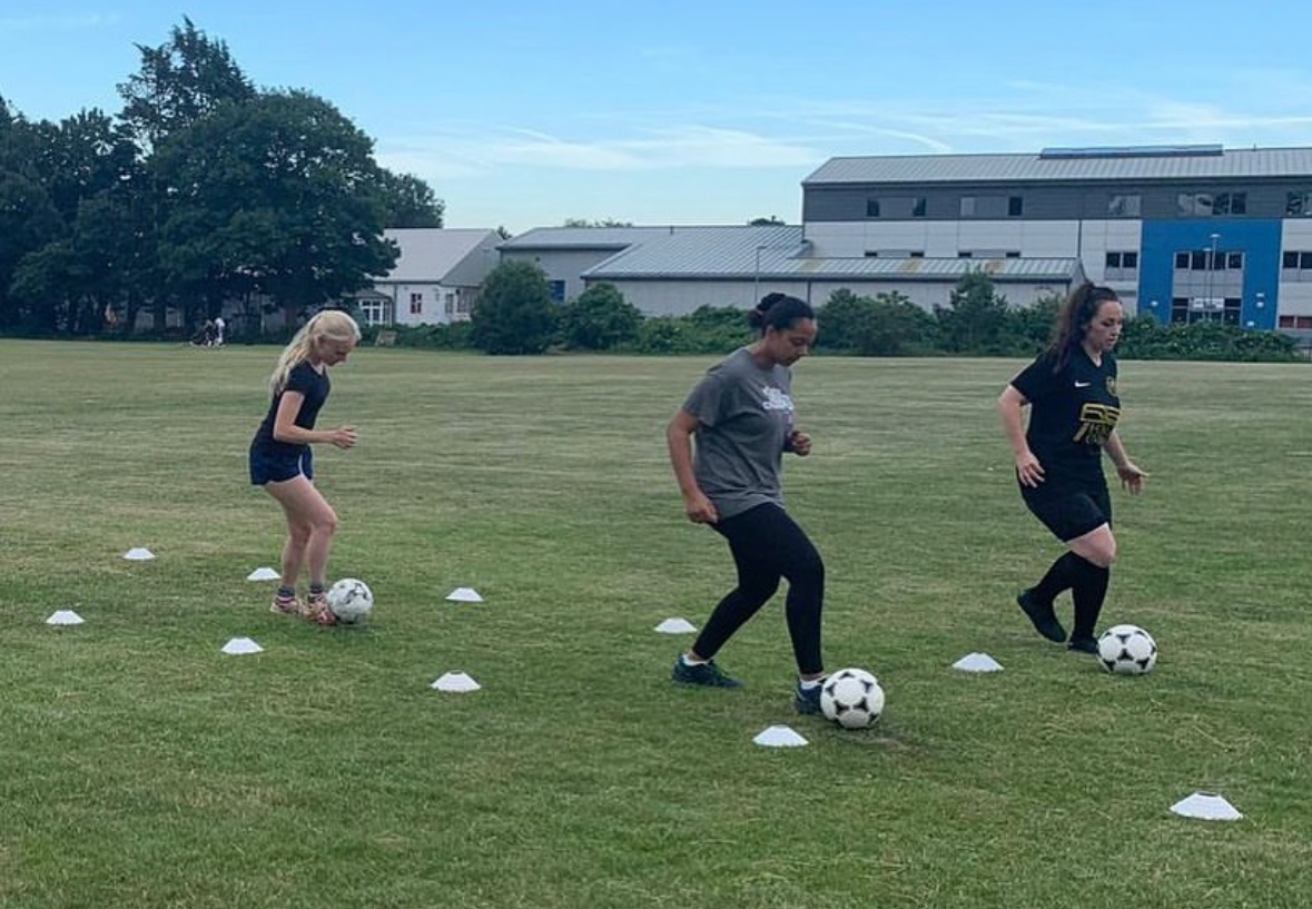 Three girls practicing dribbling on a grass field with white cones.