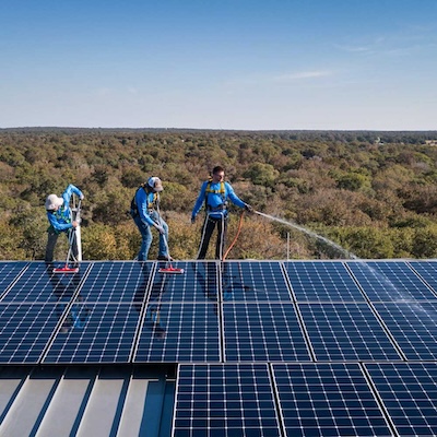 Three workers cleaning solar panels on a rooftop with a forest landscape in the background.