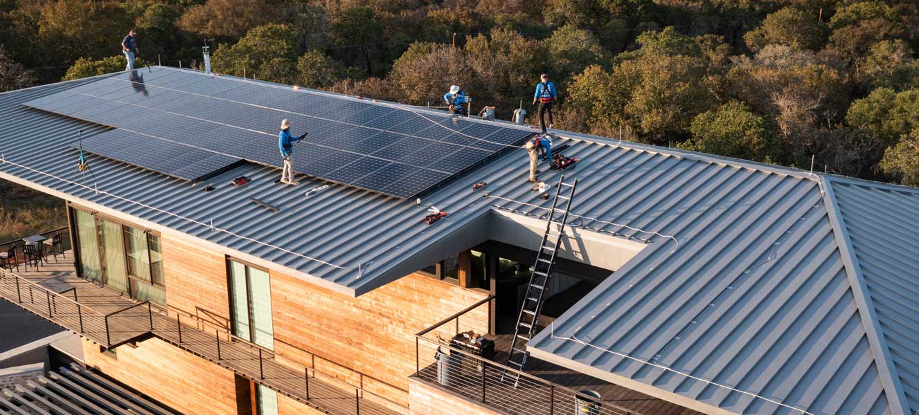 Workers installing solar panels on a metal rooftop of a modern wooden house surrounded by trees.