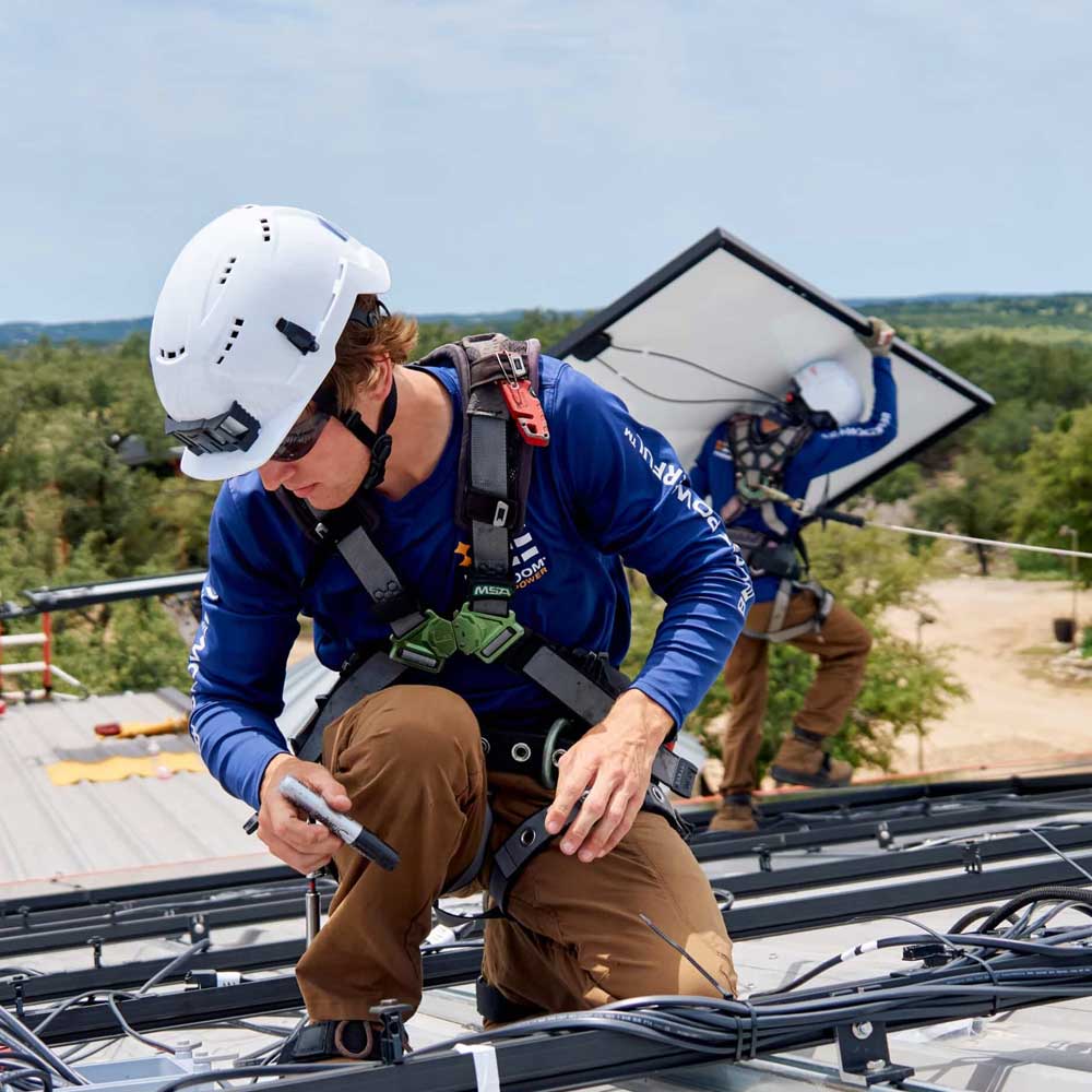 Two Freedom Power installers wearing safety harnesses and helmets installing solar panels on a rooftop with trees in the background.
