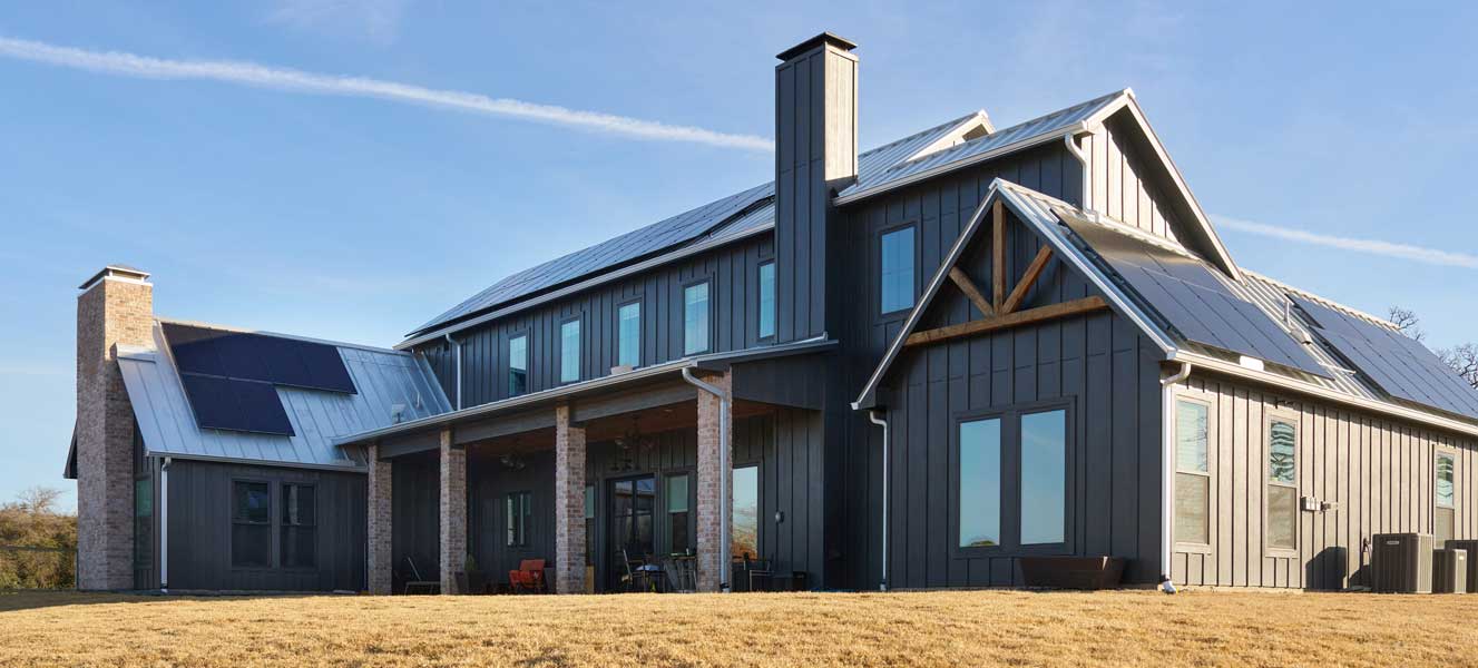 Modern farmhouse with dark siding, metal roof, and solar panels under a clear blue sky.