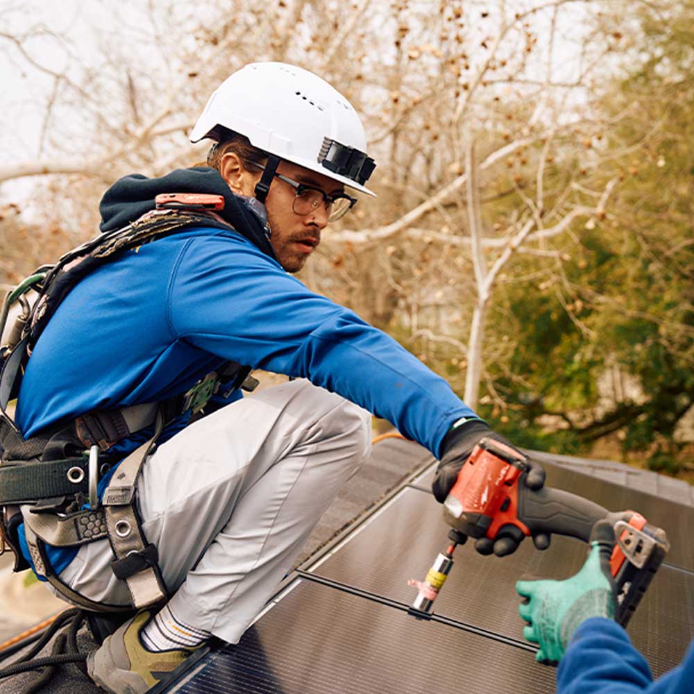 Worker in safety gear installing solar panels using a power drill on a rooftop.