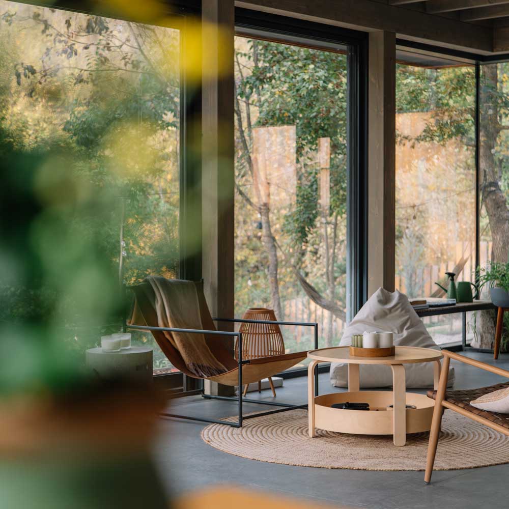 Cozy indoor seating area with modern armchairs, a round wooden coffee table, a bean bag, and large windows overlooking a forest.