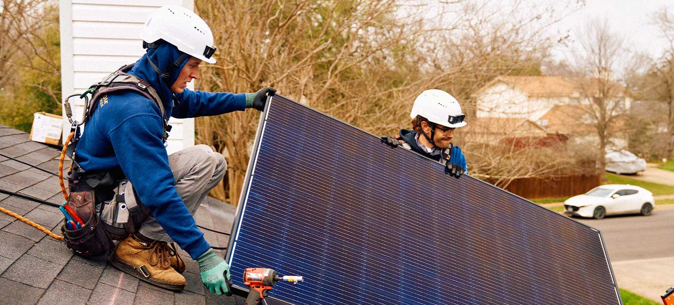 Two workers wearing helmets and safety gear install a large solar panel on a rooftop.