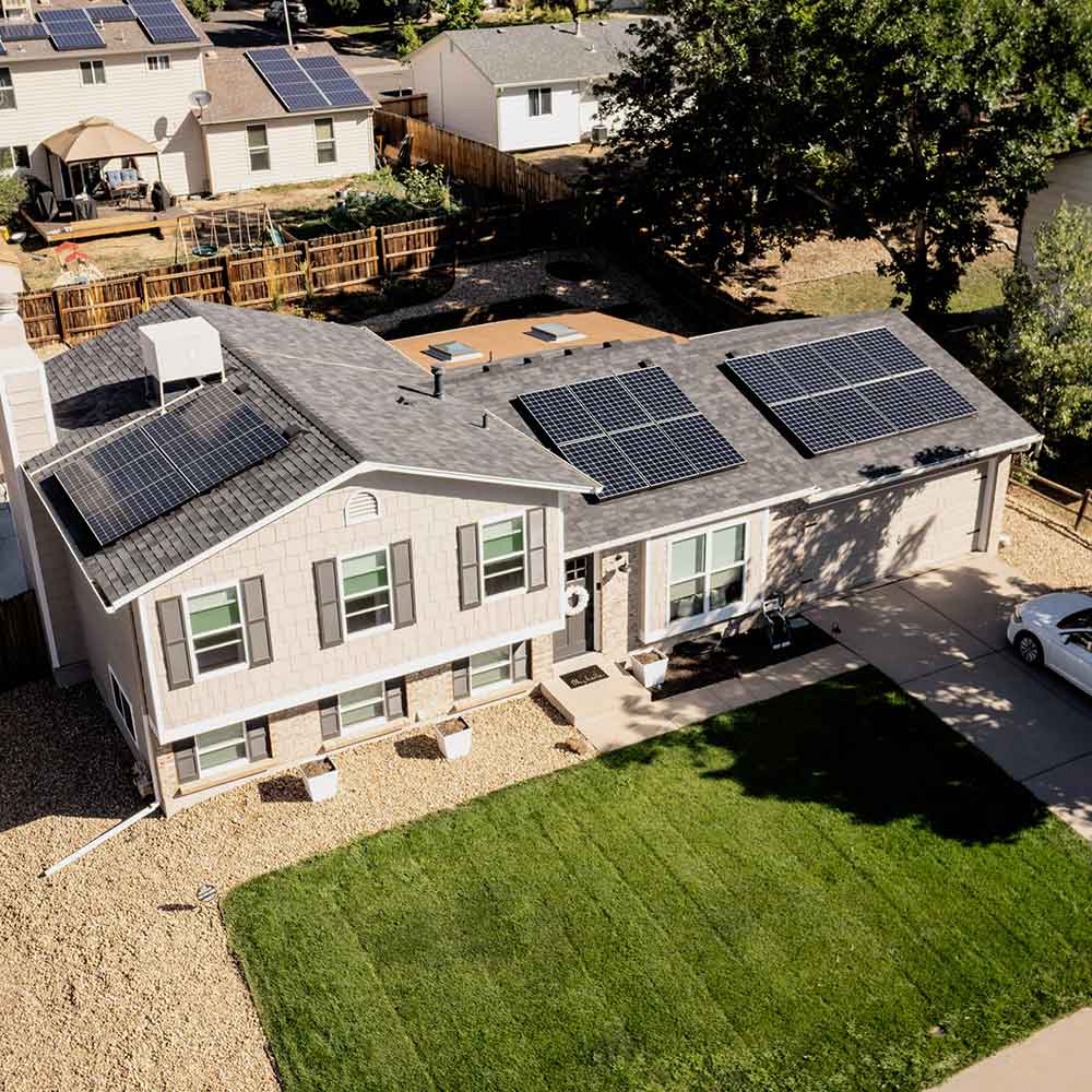 A two-story house with multiple solar panels installed on the roof, green lawn in front, and neighboring houses in the background.