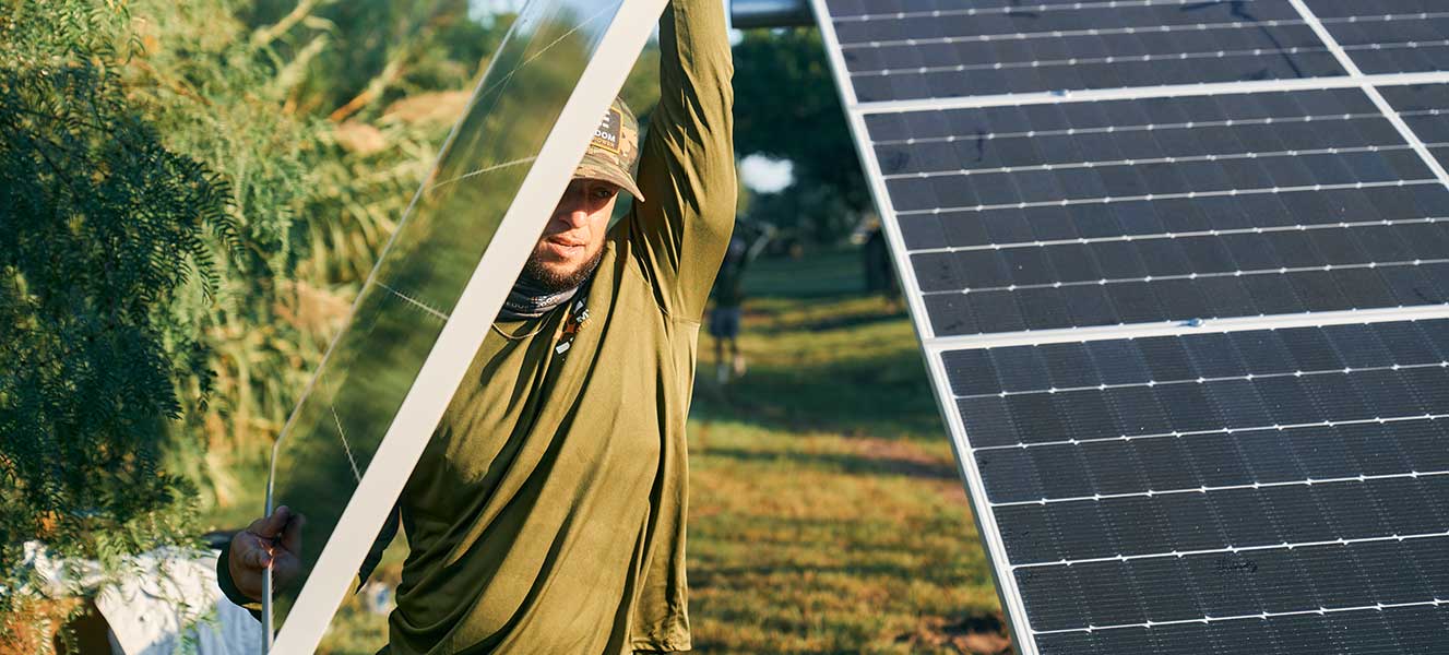 Freedom Power solar installer wearing a cap and olive green shirt installing large solar panels outdoors in a grassy area.