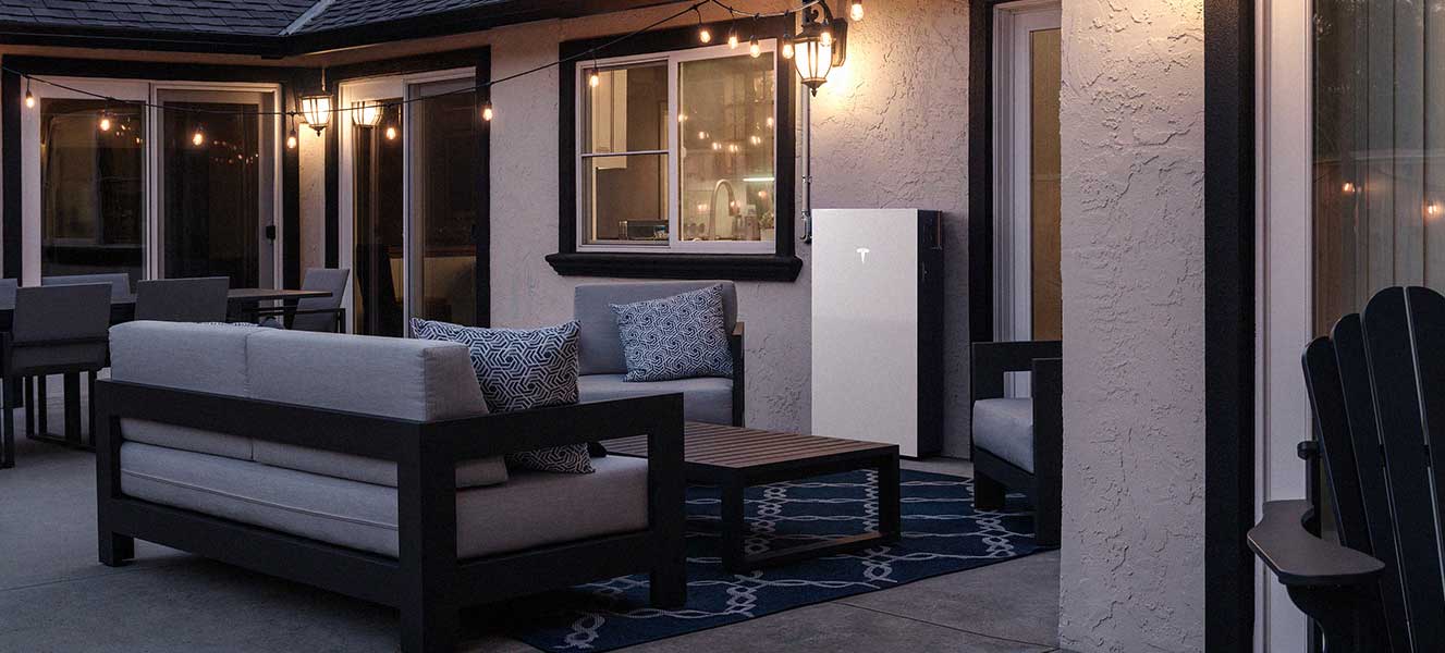 Modern outdoor patio at dusk with grey cushioned seating, a wooden coffee table, and a white Tesla Powerwall mounted on the house wall.