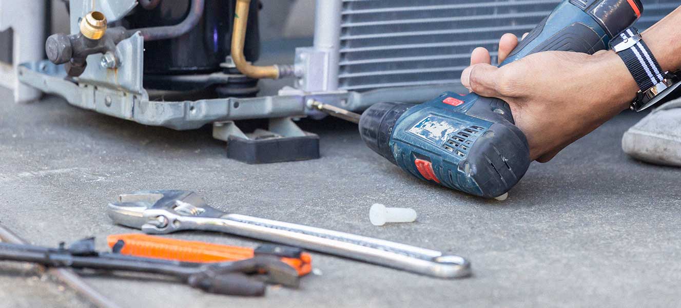 Person using a cordless drill to repair a metal appliance with a wrench, utility knife, and bolt on the floor.