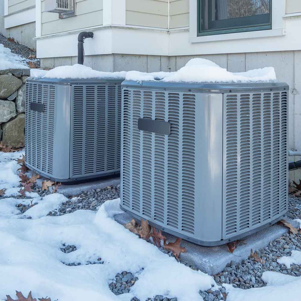 Two outdoor HVAC units covered with snow next to a house during winter.