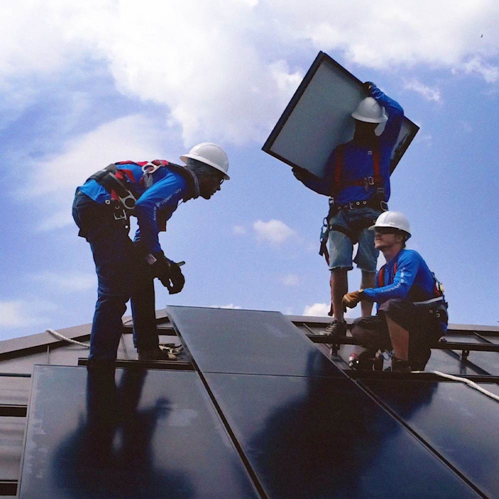 Three Freedom Power solar installers wearing safety harnesses and helmets installing solar panels on a rooftop under a partly cloudy sky.