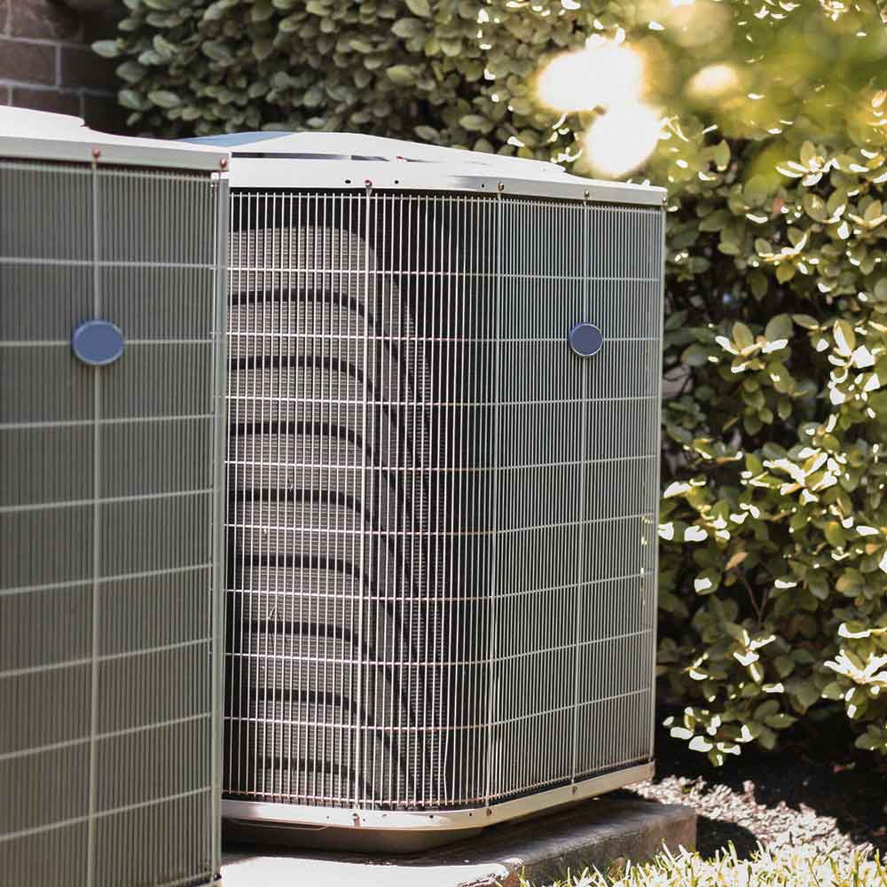 Outdoor HVAC air conditioning units beside a brick wall and leafy bushes in sunlight.