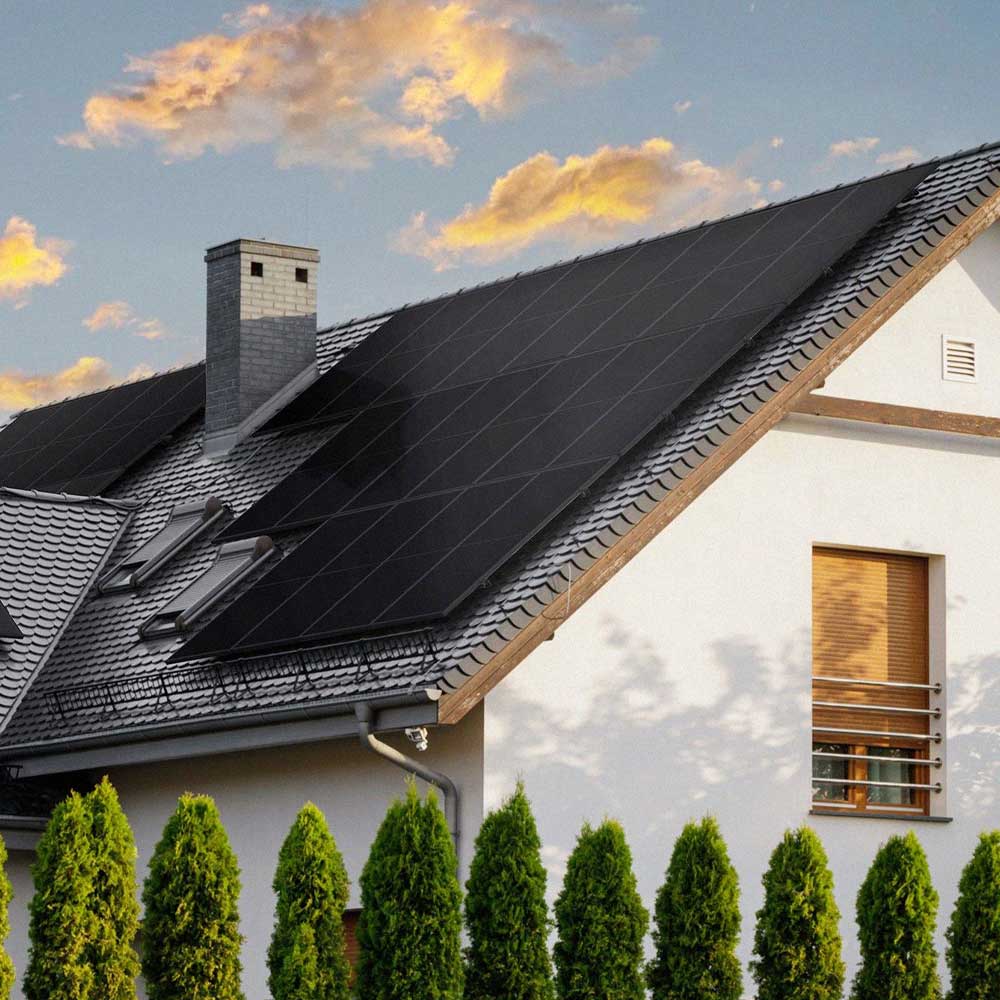House with black solar panels installed on a gray tiled roof, with a chimney and a row of green trees in front under a partly cloudy sky at sunset.
