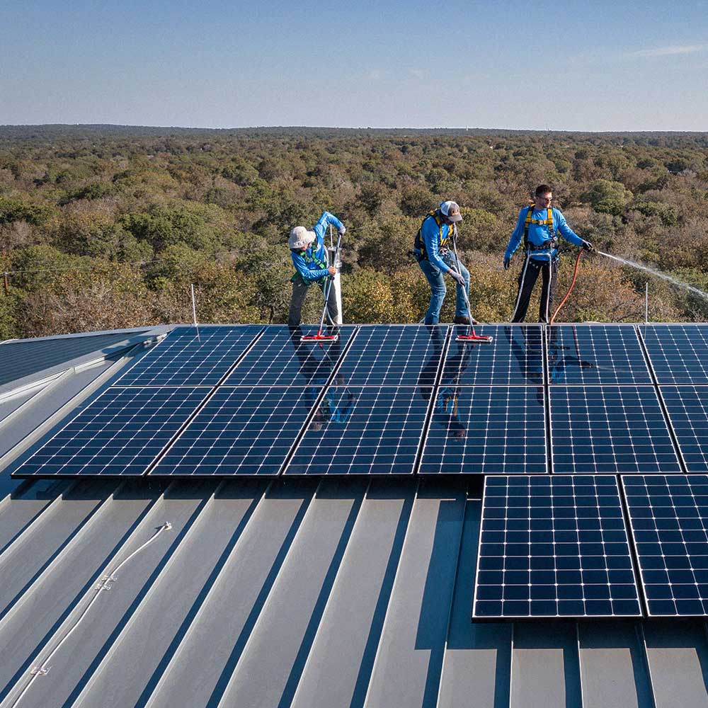 Three Freedom Power installers cleaning solar panels on a metal roof with a forest in the background under clear sky.